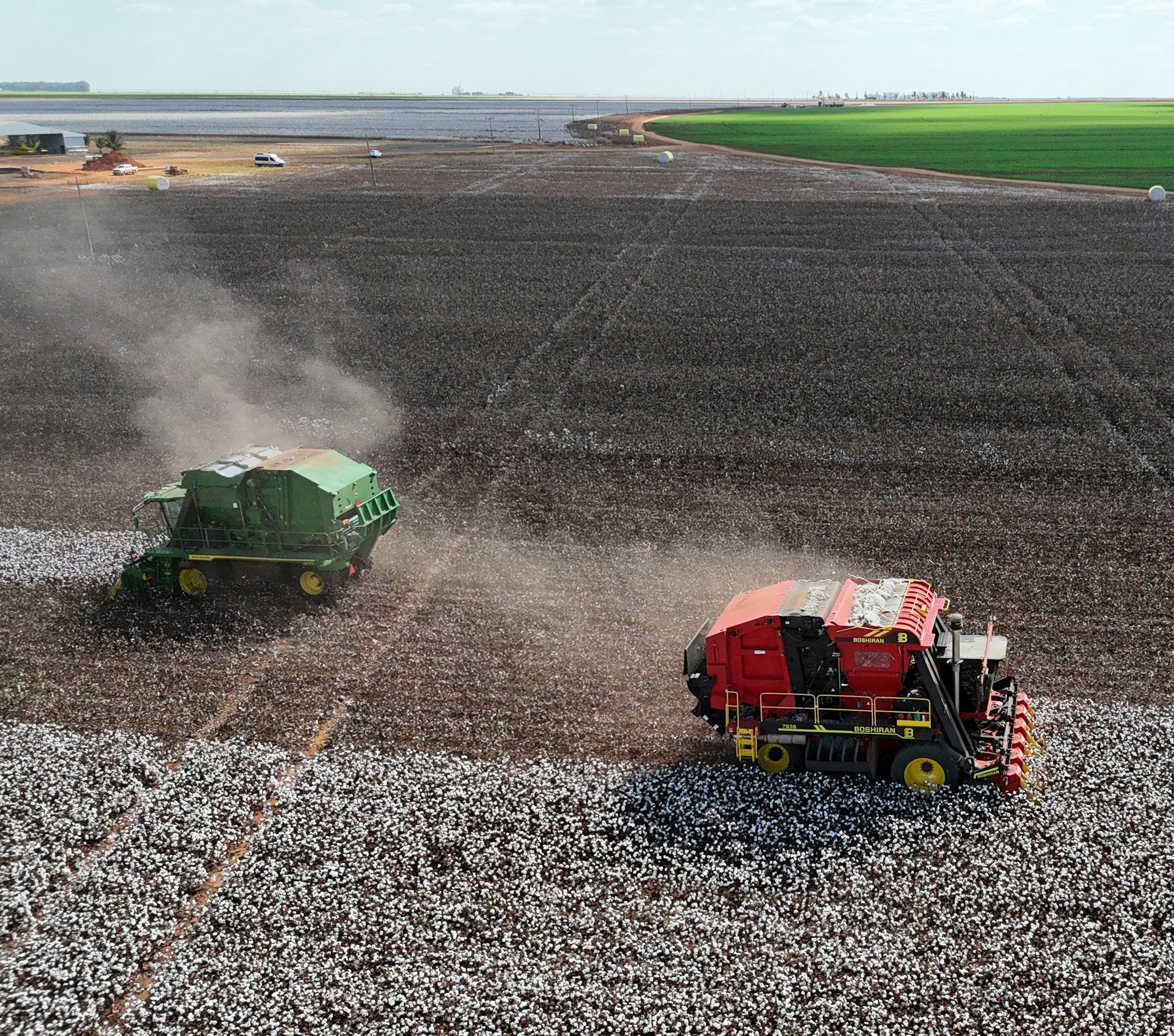 Two agricultural machines working on a field with crops and a water body in the background.