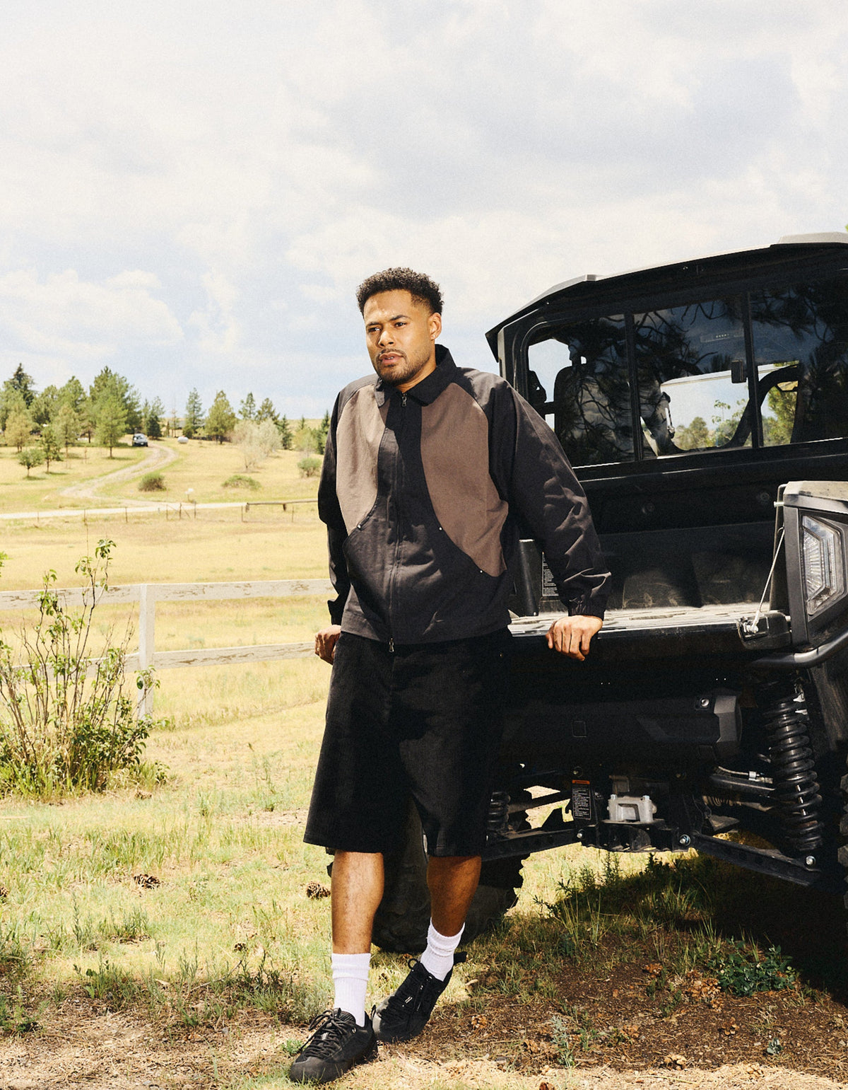 A man stands outdoors next to a black off-road vehicle in a grassy field under a cloudy sky, wearing a dark zip-front jacket, black shorts, white socks, and black sneakers.