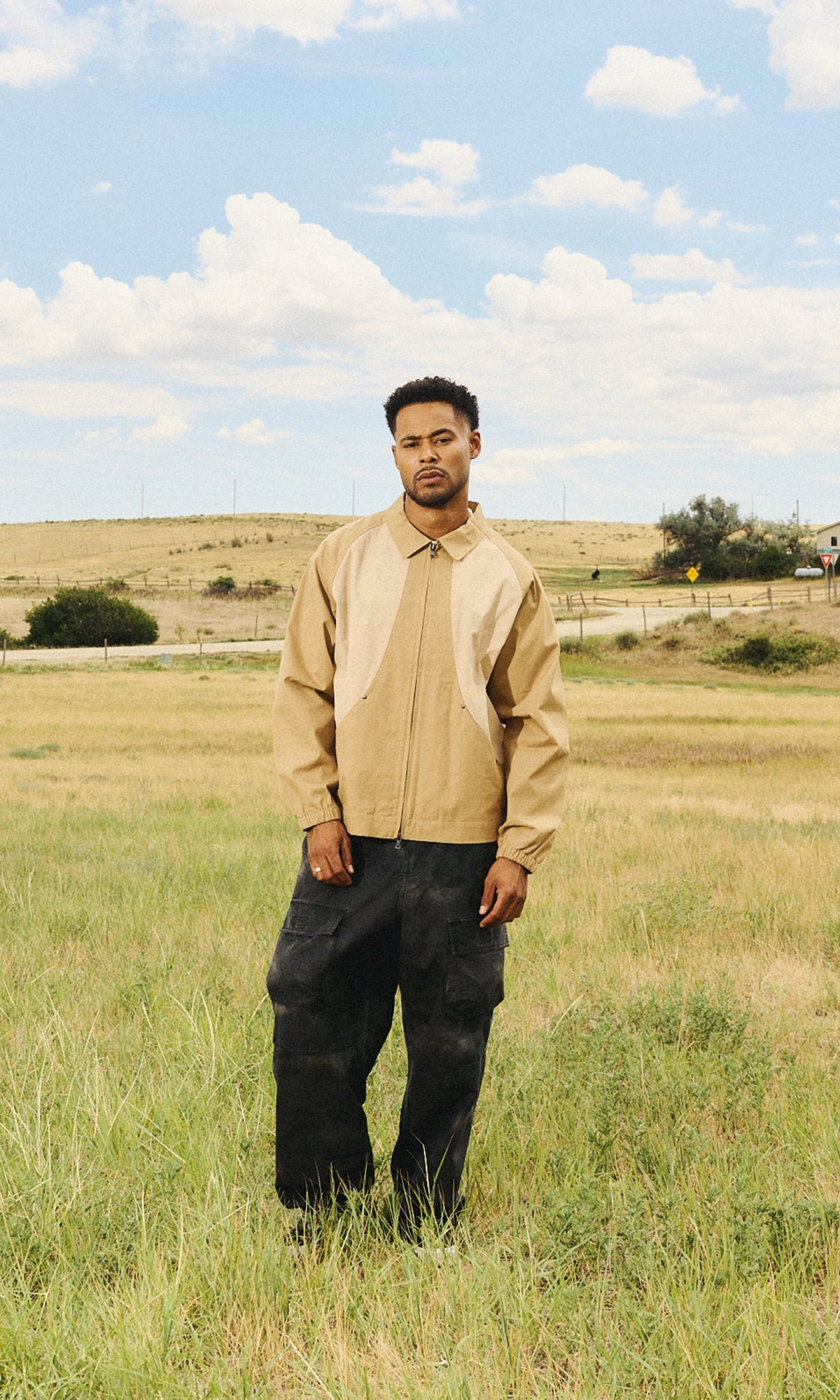 A man stands in a grassy field under a blue sky with clouds, wearing a beige zip-front jacket with white side panels and dark pants.