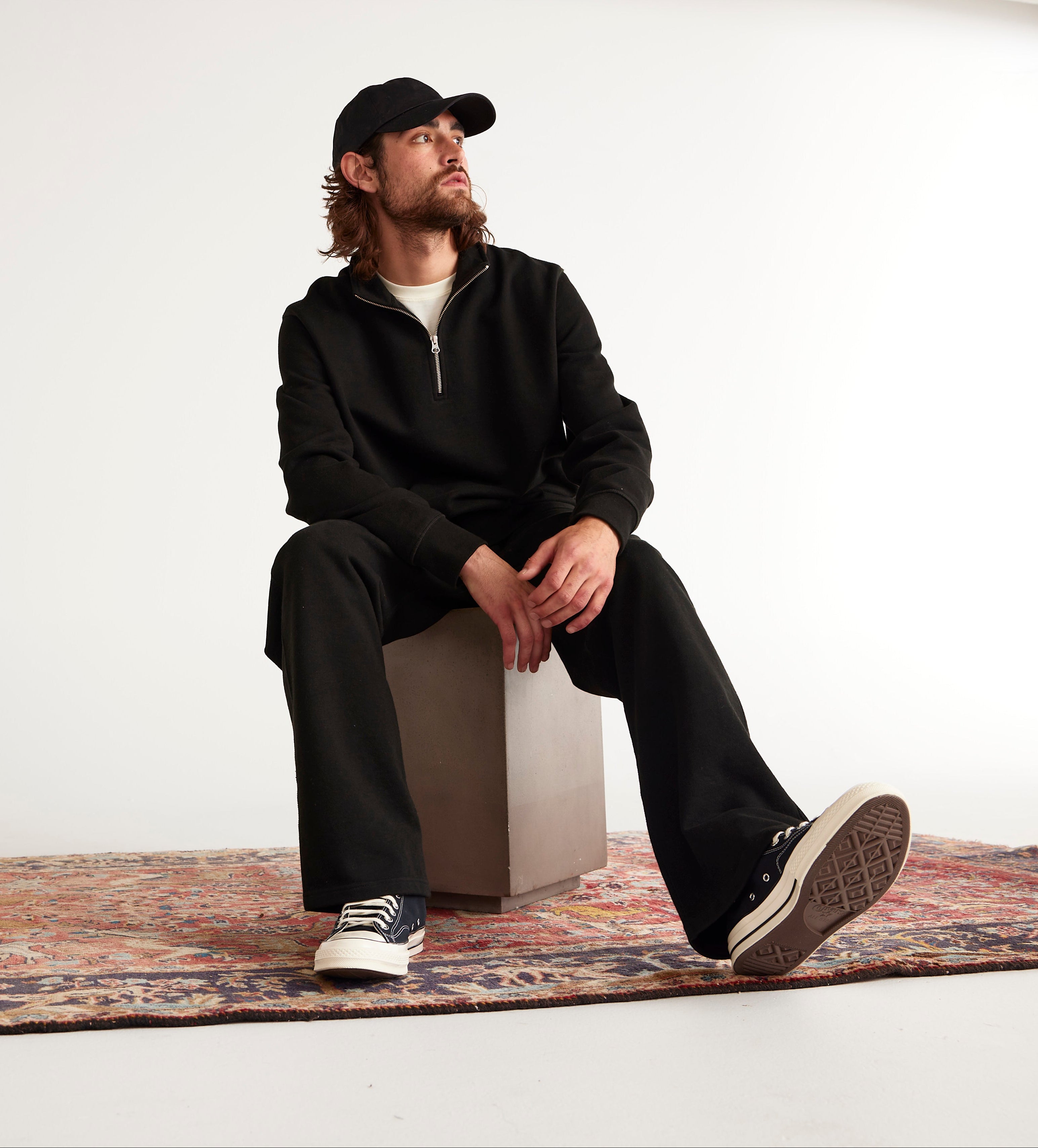 A man wearing a black zip-up jacket, black pants, a black baseball cap, and black-and-white sneakers sits on a concrete block atop a patterned rug in a minimalist studio setting.