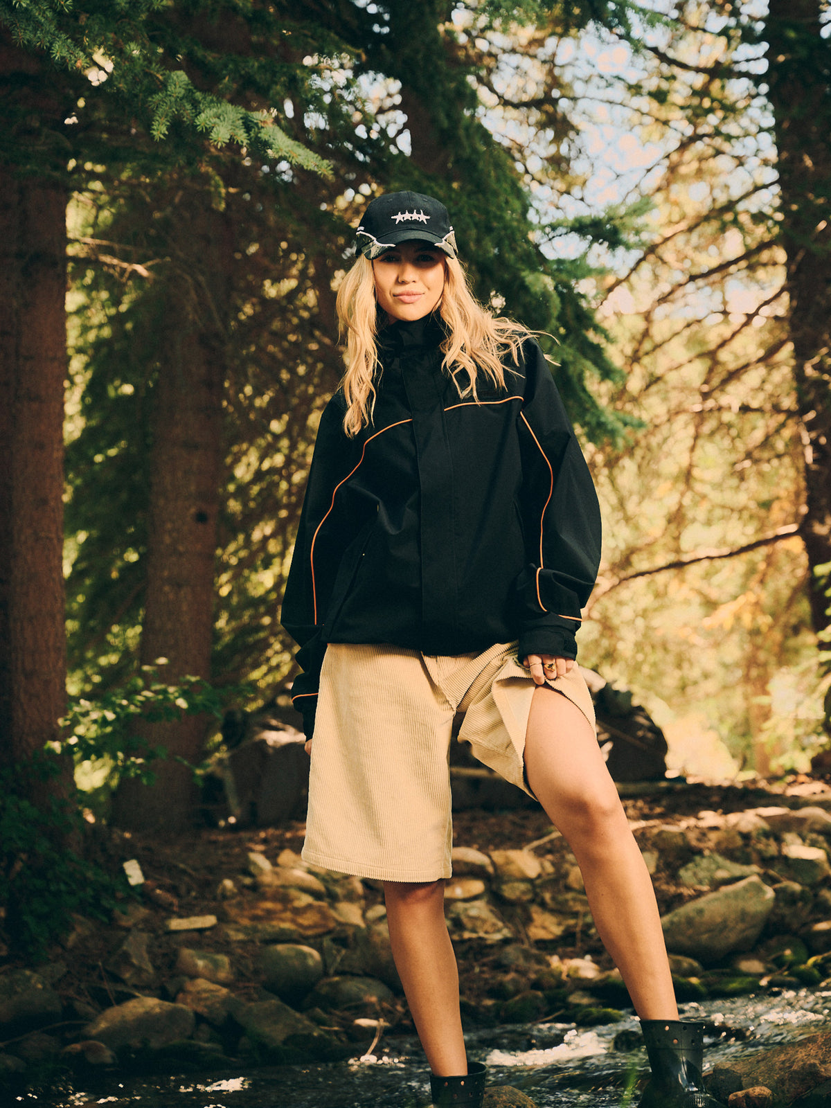 A woman stands in a rocky forest stream wearing a black lightweight jacket with orange piping, a beige mini skirt, black boots, and a black baseball cap.