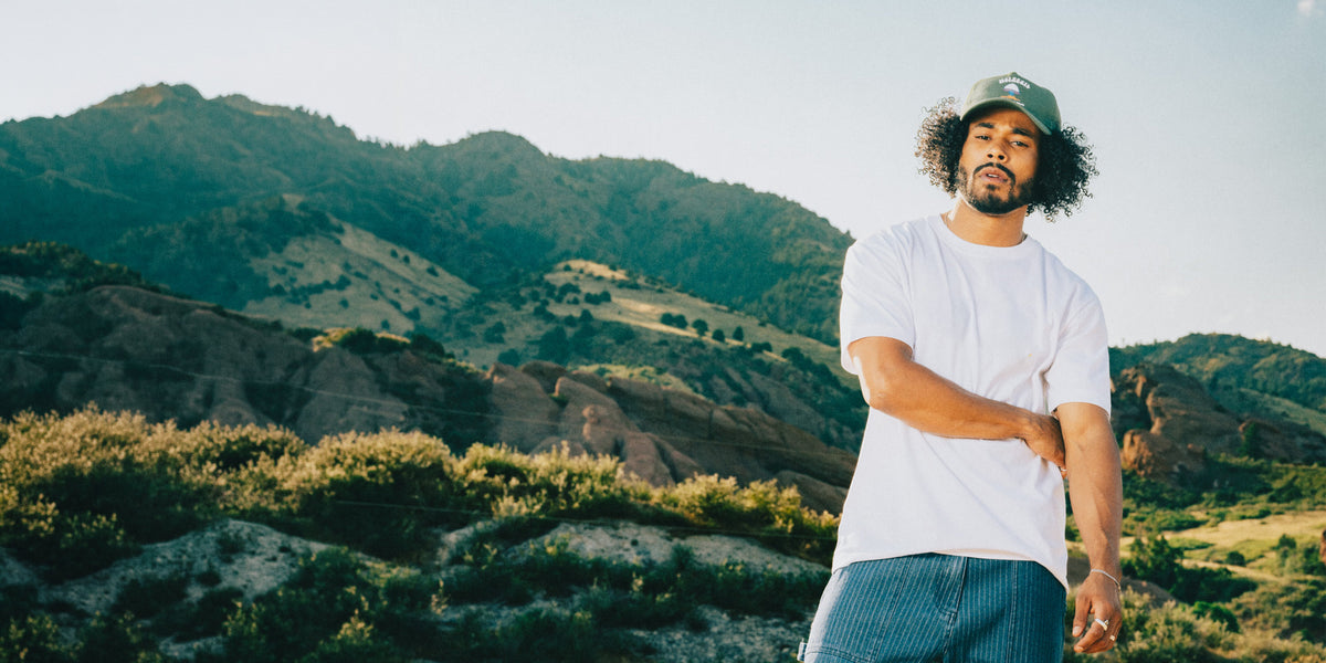 A man wearing a white t-shirt, blue striped shorts, and a green cap stands outdoors in front of a mountain landscape, looking directly at the camera.