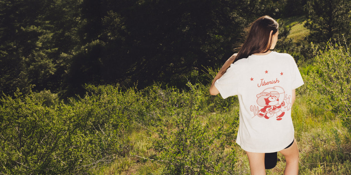 A woman stands outdoors in a grassy, wooded area, viewed from behind, wearing a white graphic T-shirt with a red cartoon character printed on the back.