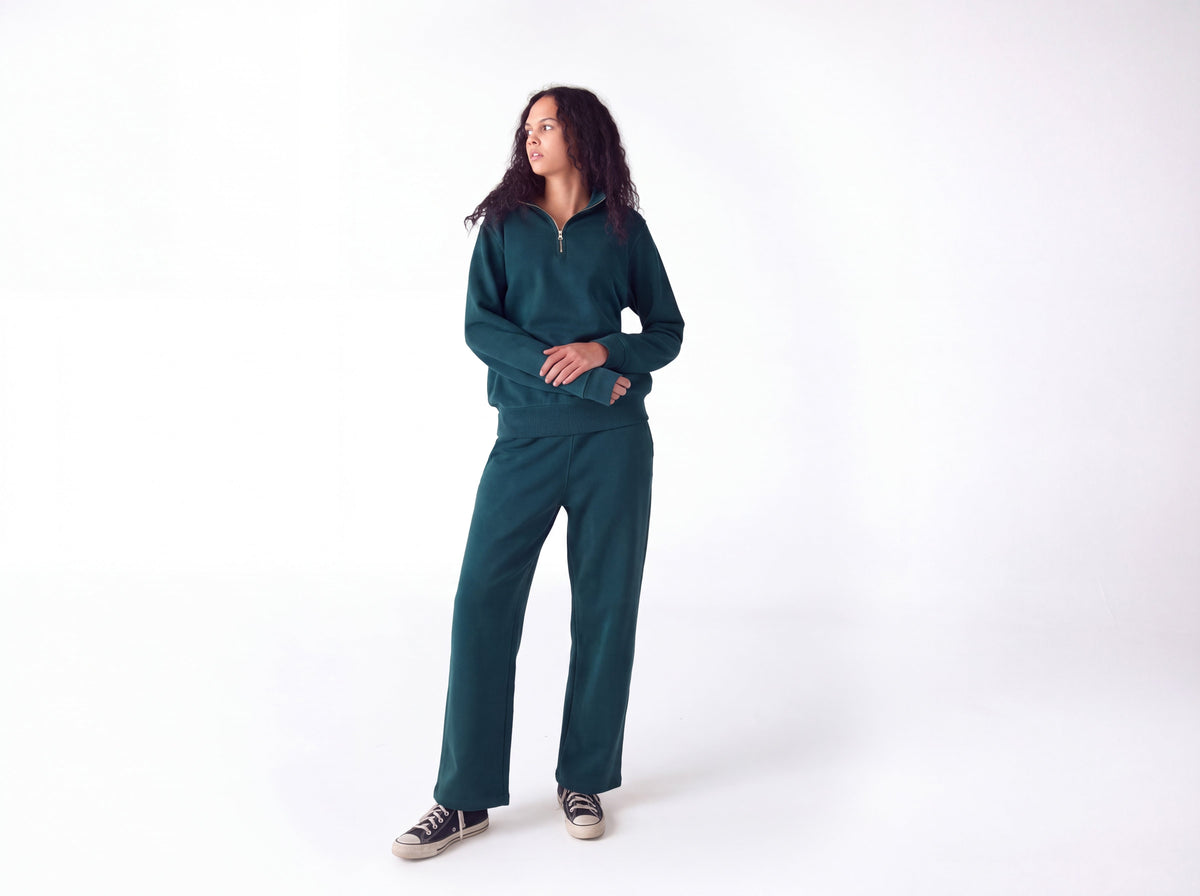 A woman modeling a matching dark teal green tracksuit, consisting of a half-zip long-sleeve pullover and wide-leg sweatpants, paired with black and white sneakers, against a plain white studio background.