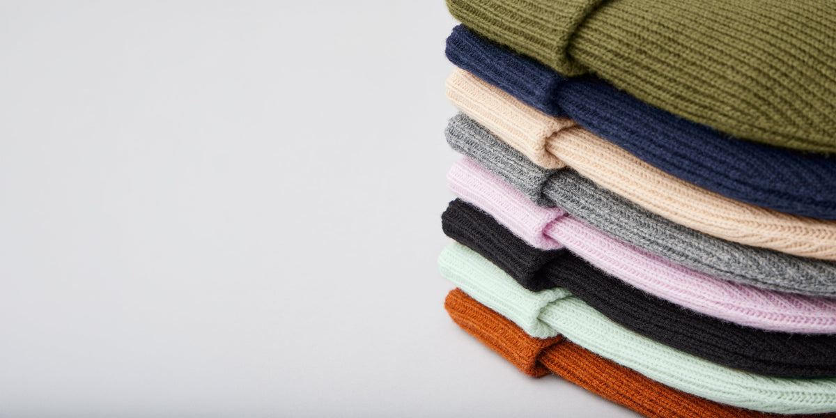 A stack of folded ribbed knit beanie hats in multiple colors, including olive green, navy, beige, gray, pink, black, mint, and rust orange, arranged on a light gray background.